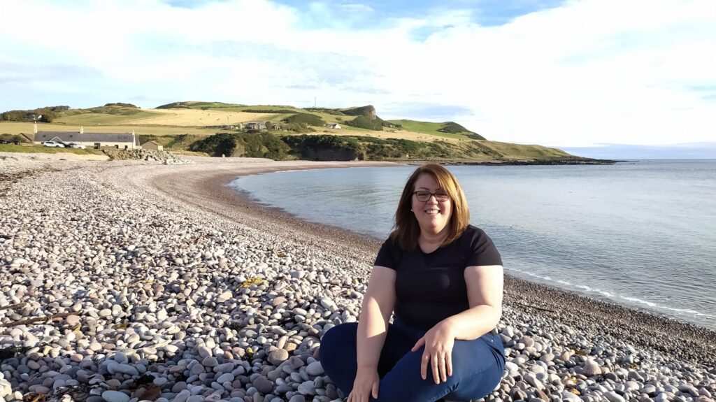 A woman is sitting on a pebbled beach with grassy hills in the background and the calm sea to her right. She is smiling and wearing a black shirt and blue jeans, embodying pure joy and love for the serene moment.