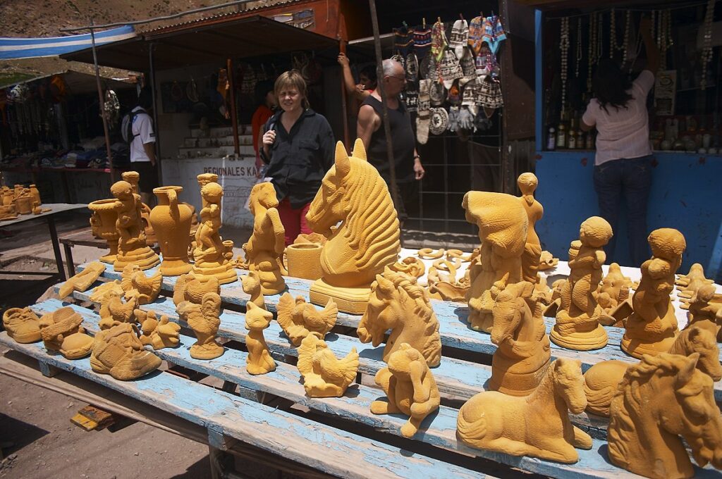 Outdoor market stall displaying various clay sculptures, including animals and decorative pieces, on wooden tables with shoppers browsing in the background.