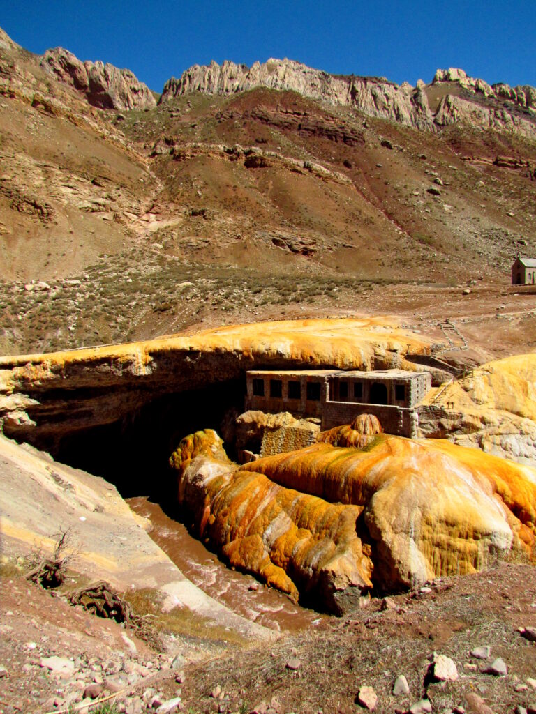 A dilapidated stone structure surrounded by multicolored mineral deposits is situated in a rugged, mountainous landscape under a clear blue sky.