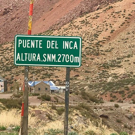 A green sign reads "Puente del Inca Altura SNM 2700m" against a rocky hillside background with a small building and sparse vegetation.