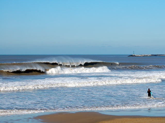 A person in a wetsuit stands on a sandy beach near the water's edge, looking out at large ocean waves under a clear blue sky, contemplating their origin.