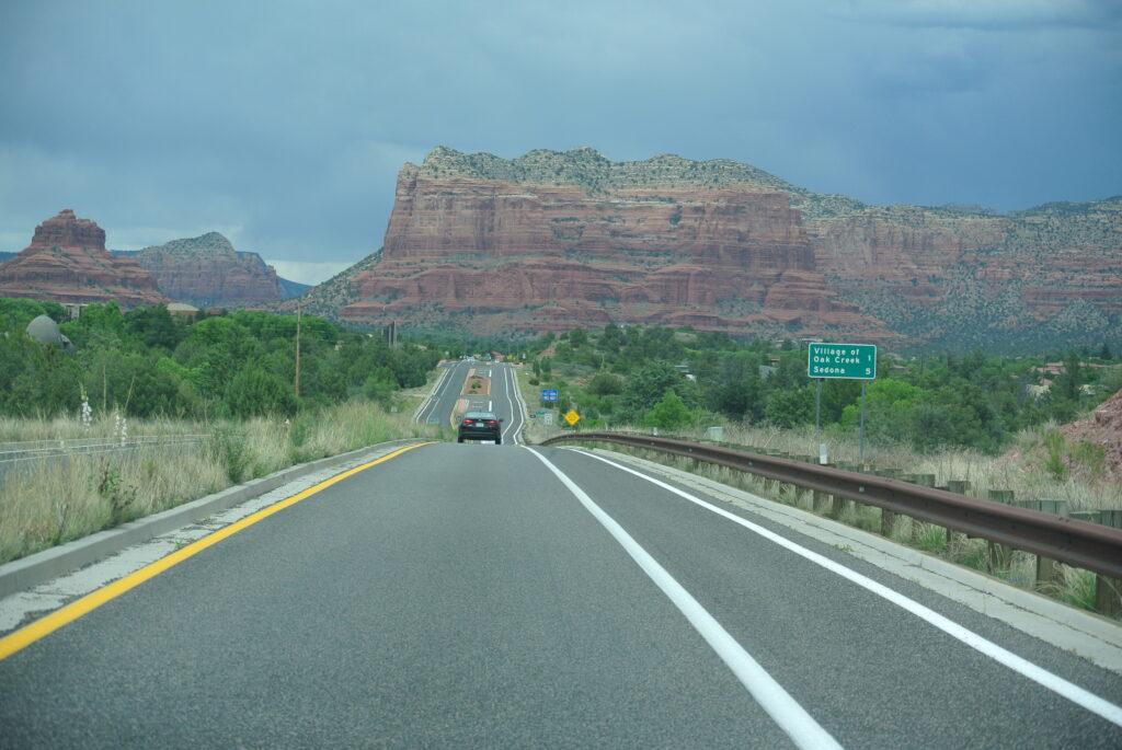 A car drives on a highway towards Sedona's red rock formations under a cloudy sky, with green vegetation on either side and a road sign visible to the right.