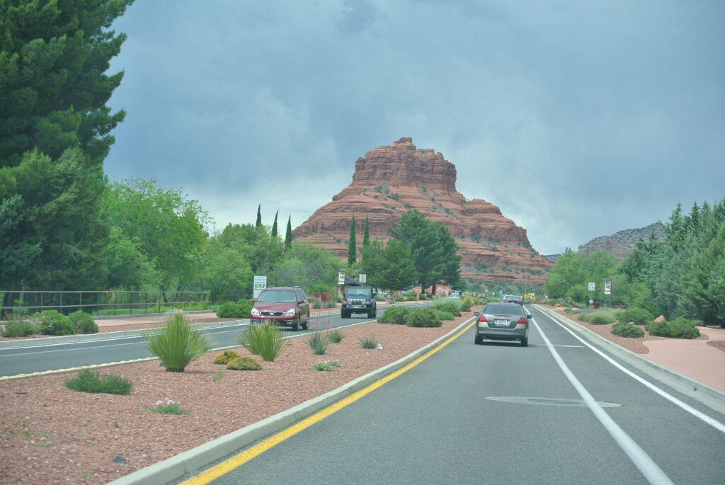 A road with cars driving closer towards a red-rock formation in Sedona, surrounded by greenery and desert plants under a cloudy sky.