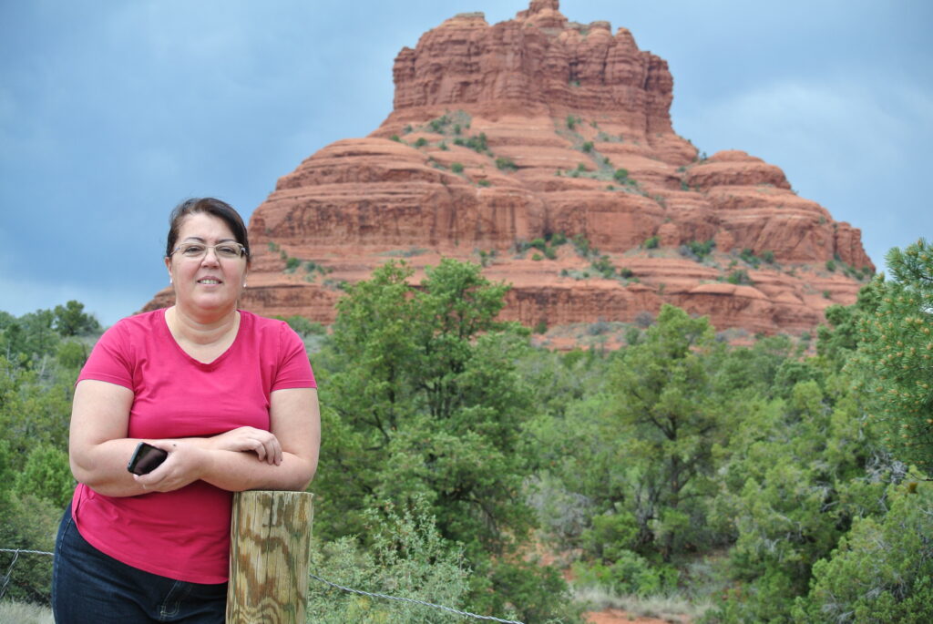 A woman in a pink shirt and jeans stands resting on a wooden post closer to the majestic red rock butte of Sedona, surrounded by green foliage under a cloudy sky.