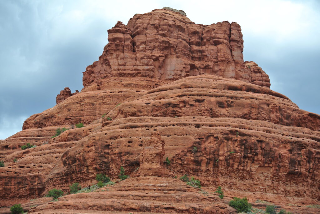 A large red rock formation with layered, textured surfaces and small scattered greenery at the base stands against a cloudy sky, reminiscent of Sedona's striking landscapes.