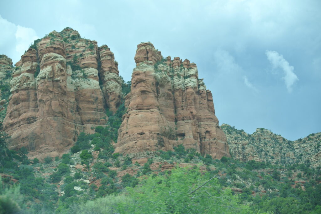 A natural rock formation with reddish, layered cliffs and green vegetation in the foreground, set against a cloudy sky; it feels like you're closer to Sedona's breathtaking beauty and stars.