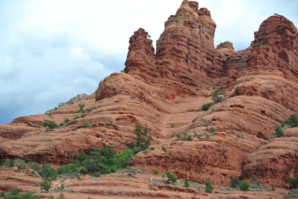 A rugged red rock formation with sparse vegetation stands under a cloudy sky in Sedona, bringing you closer to nature's raw beauty.
