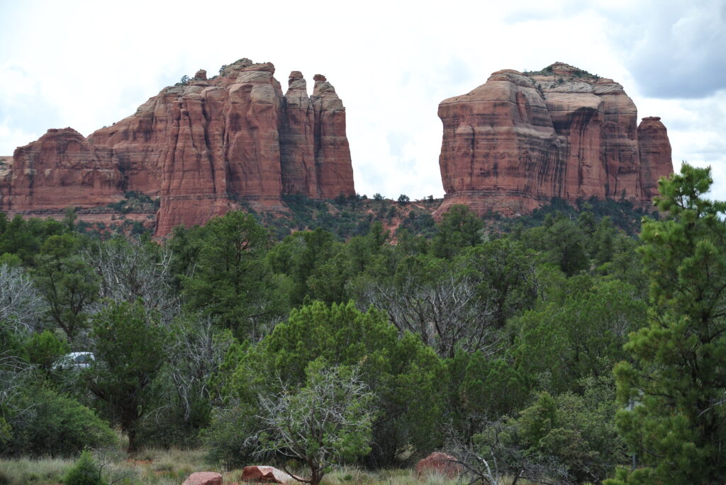 A landscape featuring tall red rock formations surrounded by green vegetation and trees under a cloudy sky, reminiscent of Sedona, brings you closer to nature.