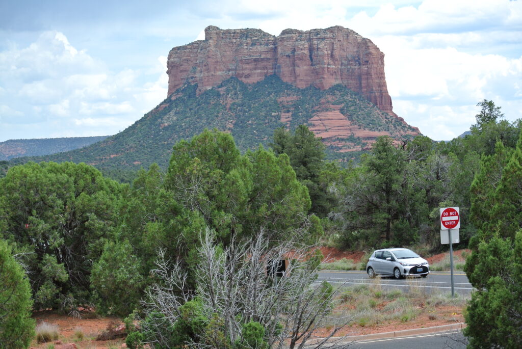 A gray car drives on a road with a "Do Not Enter" sign ahead, surrounded by green shrubs and trees. In the background, closer to the horizon, there is a large red rock formation under a partly cloudy sky, reminiscent of Sedona's stunning landscapes.
