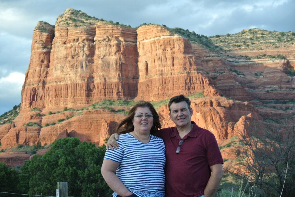 A woman and a man stand together outdoors in Sedona with their arms around each other, smiling at the camera. Behind them, the large red rock formation with layers and green vegetation at the base draws you closer to nature's artwork under a canopy of stars.