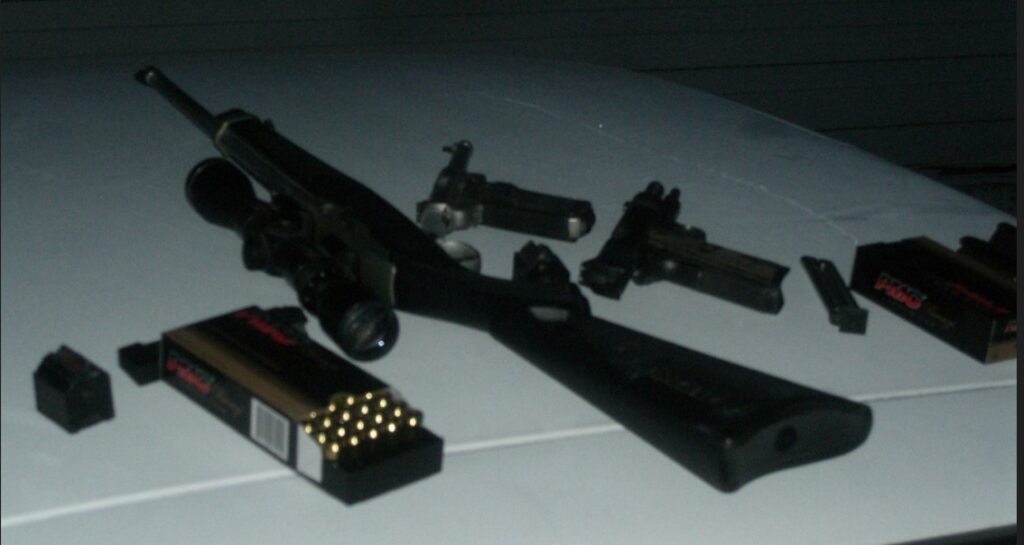 A rifle with a scope, two handguns, ammunition, and two boxes of bullets are displayed on a flat surface, reminiscent of the arsenal Bonnie and Clyde might have carried.