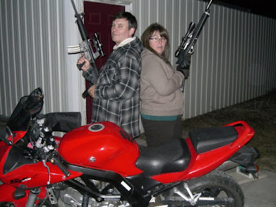 Two individuals stand back-to-back, channeling a modern Bonnie and Clyde, holding rifles next to a red motorcycle outdoors. A metal building and a door are visible in the background.
