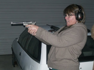 A woman wearing earmuffs aims a handgun at an indoor shooting range, evoking the spirit of Bonnie behind a white car.
