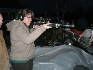 A person wearing glasses and earmuffs aims a rifle outdoors at night while another stands nearby, reminiscent of a modern-day Bonnie and Clyde. A motorcycle can be seen in the background.
