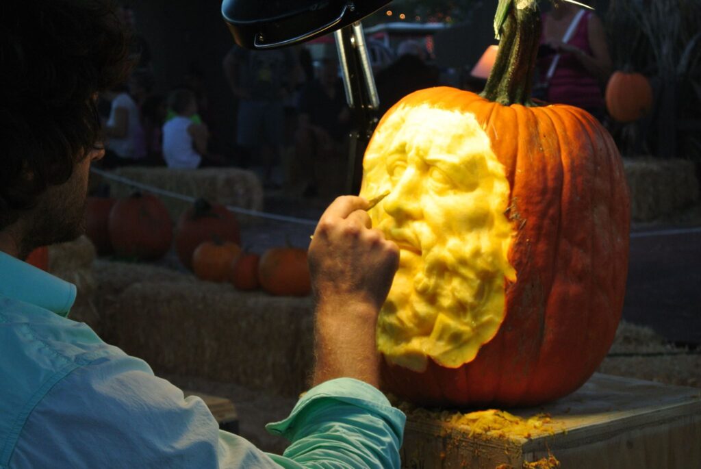 A skilled artist carves a detailed human face into a large pumpkin under focused lighting, with other pumpkins and hay bales adding to the festive backdrop.
