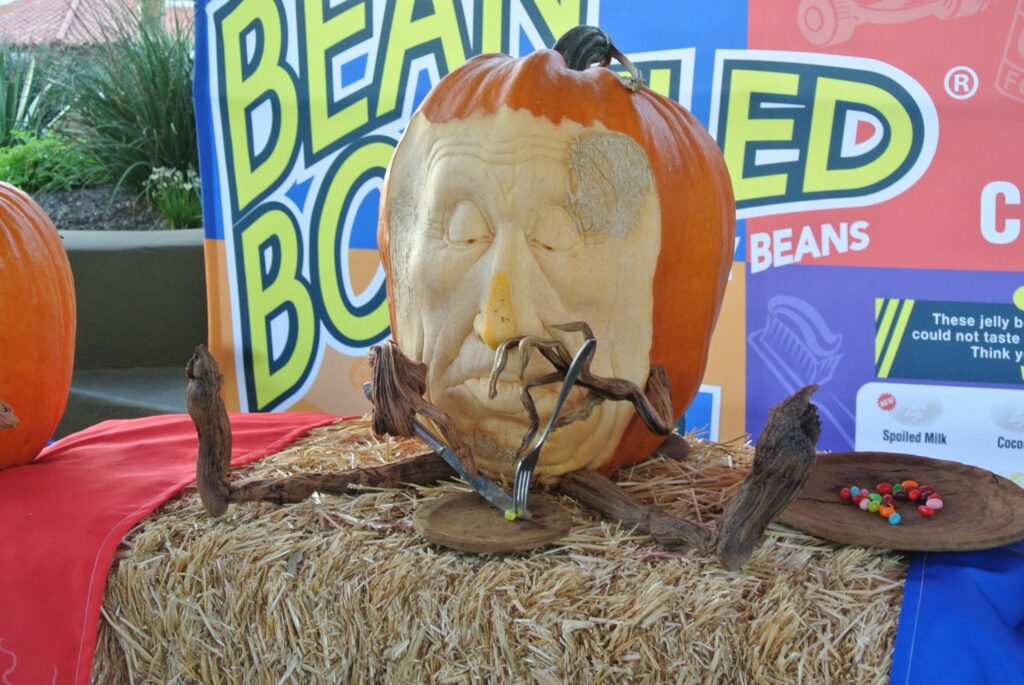 A carved pumpkin with a human face, glasses, and wooden arms sits on a hay bale. Surrounding it, signs and colorful backgrounds with other pumpkins are partially visible.
