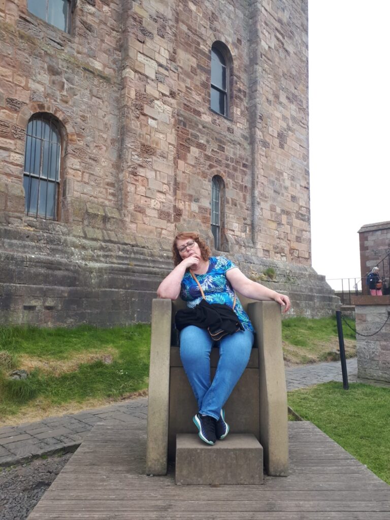 A woman in casual clothing sits on a stone chair in front of an old stone building with barred windows, lost in thought.