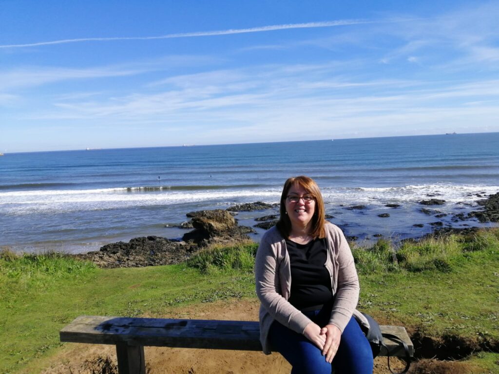 A woman is sitting on a bench by the sea, waves crashing on rocks in the background. She is smiling, wearing glasses, and the clear blue sky stretches above. It's moments like these that make her pause to think about how thankful she is for the beauty around her.