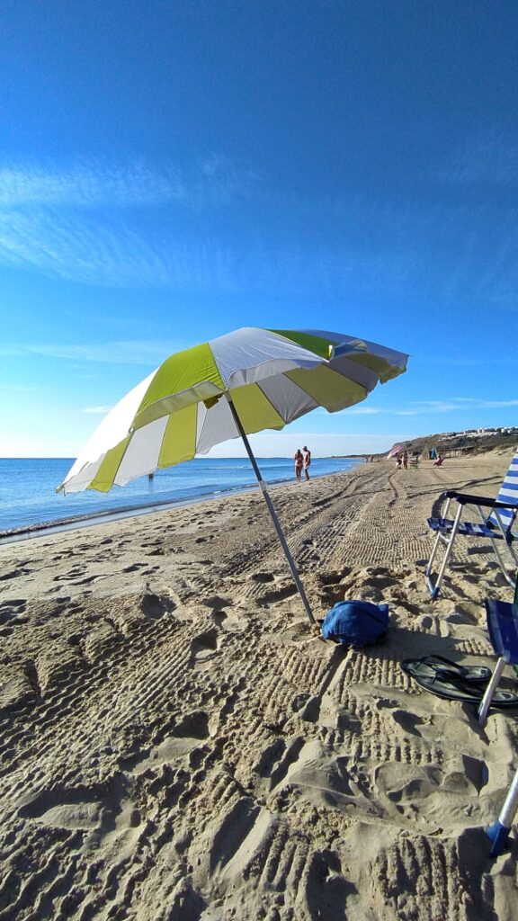 A white and green beach umbrella is set up on a sandy shore near the water's edge, with two folding chairs and a blue bag beside it. People are walking along the beach on a sunny day, full of gratitude for nature's beauty.