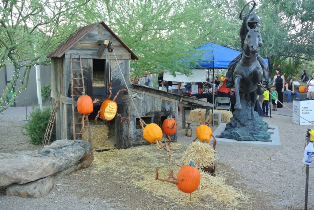 An outdoor setup features intricately carved pumpkins surrounding a wooden structure and a bronze horse statue. People in the background are gathered near a blue tent.