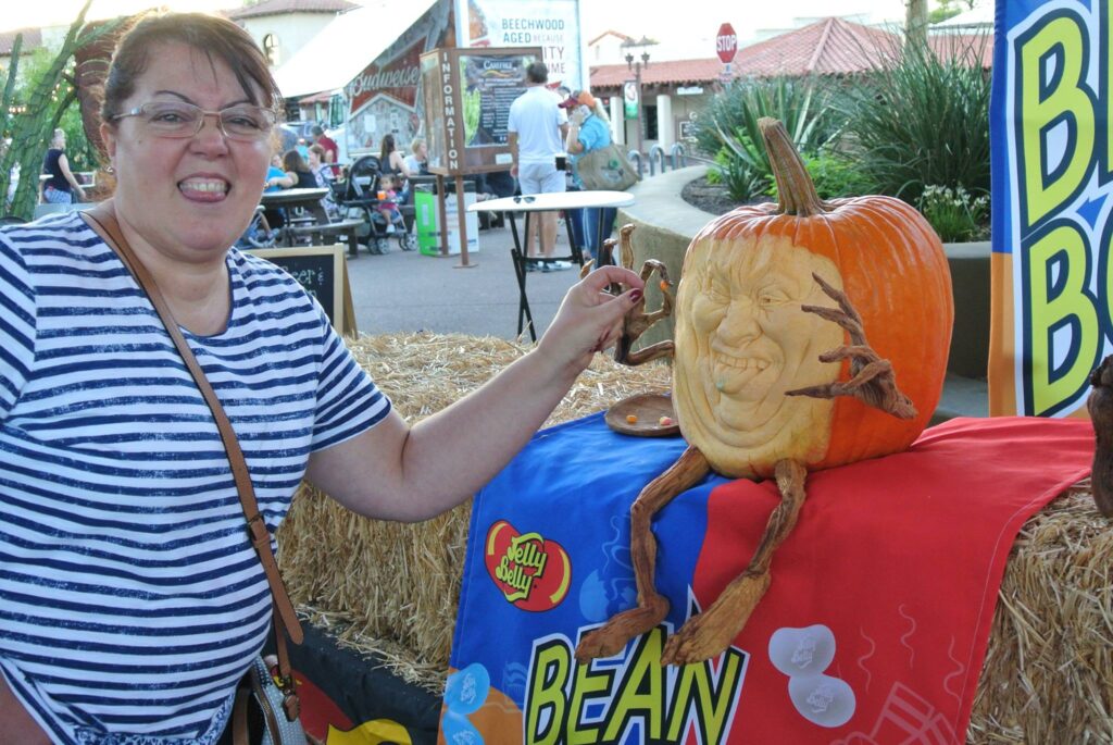 A smiling woman poses next to a carved pumpkin with arms, legs, and a face, set up on a colorful table display at an outdoor pumpkins-themed event.