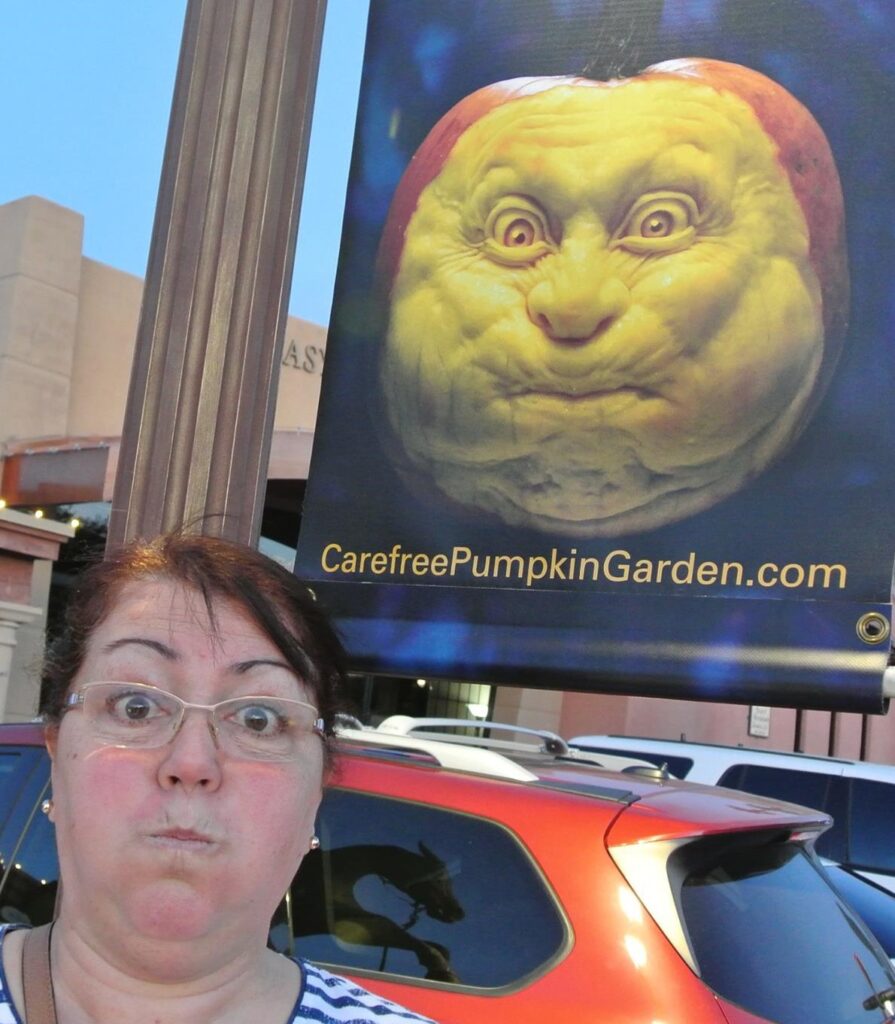 A woman with glasses mimics the comical facial expression of a carved pumpkin displayed on a sign behind her, adding to the festive spirit of pumpkins.