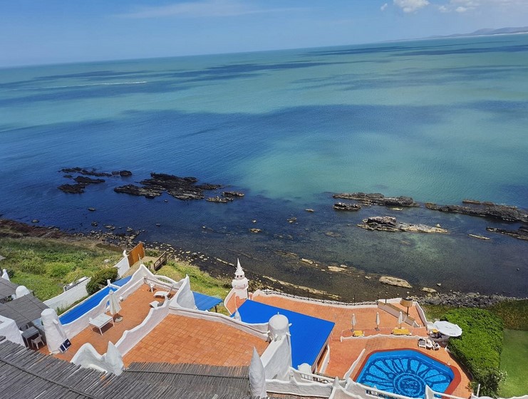 A coastal view on a magical canvas features white buildings with terracotta roofs, a circular blue swimming pool, and a rocky shoreline extending into a calm blue-green sea under a clear sky.