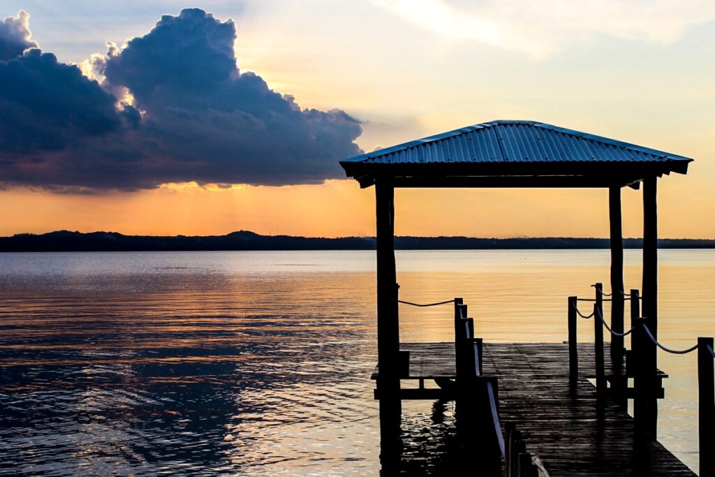 A wooden pier with a shelter extends into calm water at sunset, with a partially cloudy sky in the background.