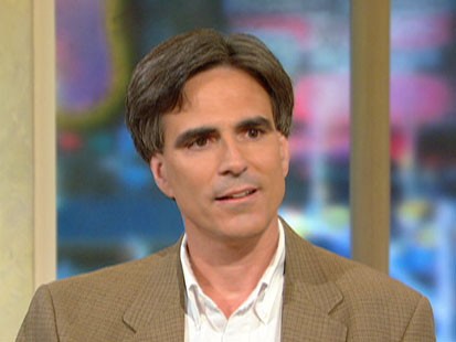 A man with short gray hair, wearing a white shirt and brown blazer, stands in front of a blurred, colorful background, contemplating a meaningful question.