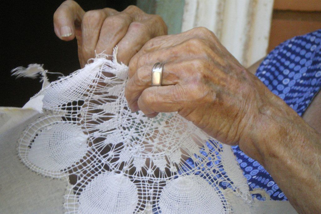 Elderly hands wearing a ring hold and work on a delicate piece of lace with intricate patterns.