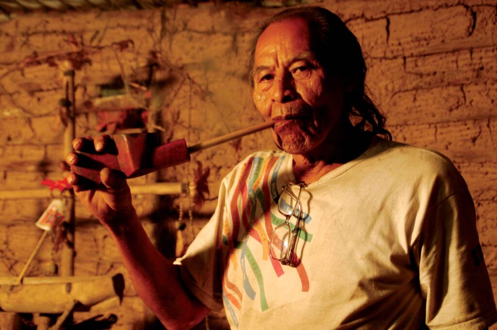 An older person with long hair and a mustache, wearing a white shirt, smokes a long pipe while standing in a rustic room with brick walls. The person has glasses hanging around their neck, resembling an enigmatic figure out of Ypacarai's folklore.