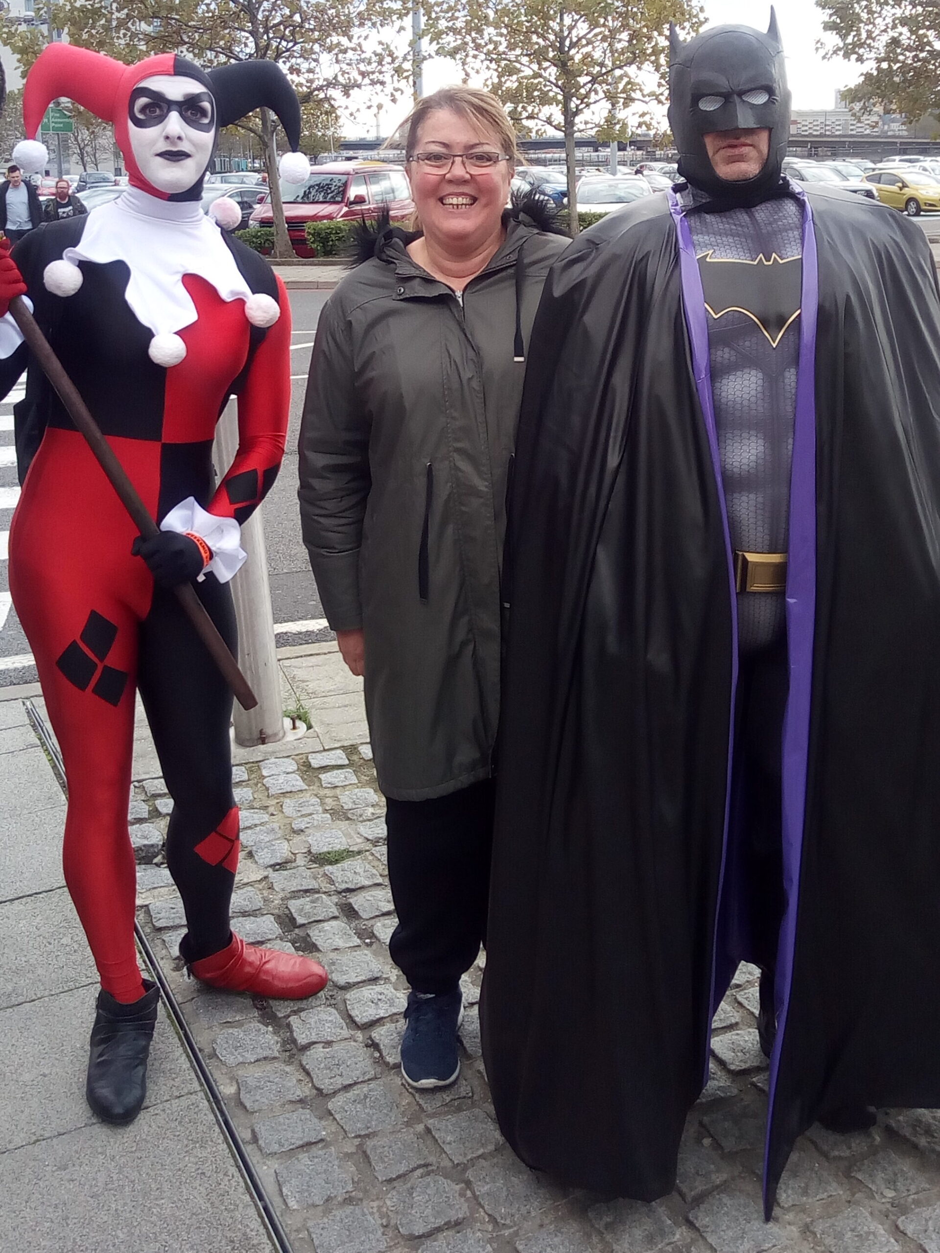 A person stands between two cosplayers dressed as comic characters, each exuding a sense of superpower, on a paved area near a street.