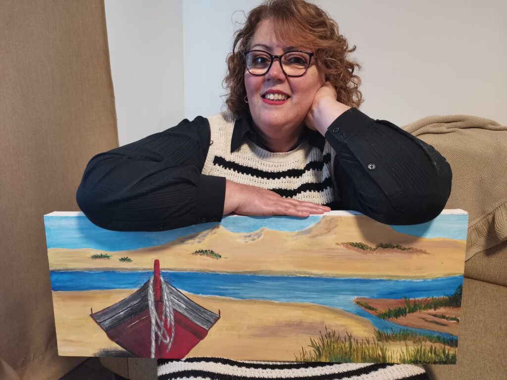 A woman smiles while sitting on a couch, displaying a painting of a red boat on a sandy shore near a blue river, sparking thoughts of adventure.