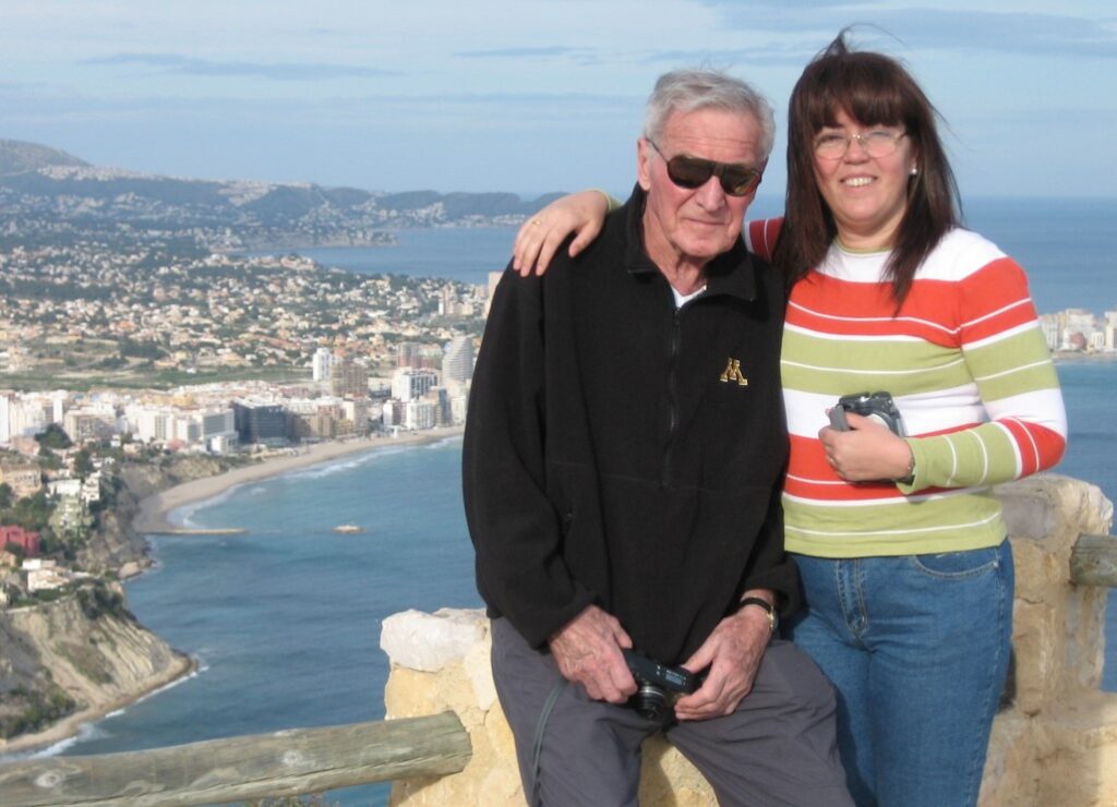 Two people stand on a scenic overlook with a coastal city and ocean in the background. The man, who became somewhat of a local legend, is wearing sunglasses, and both are holding cameras.