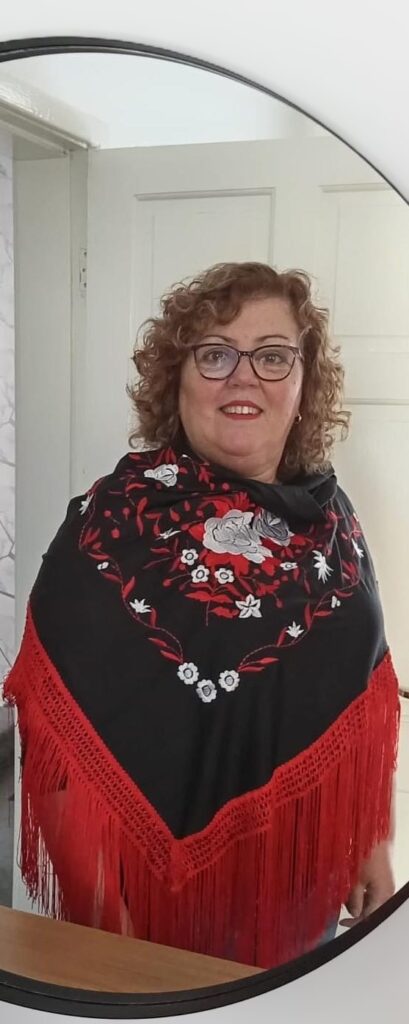 A woman with curly hair and glasses stands indoors, her black shawl adorned with red and white floral embroidery and fringes reminiscent of a vibrant fiesta, casting a lively contrast against the plain white door behind her.
