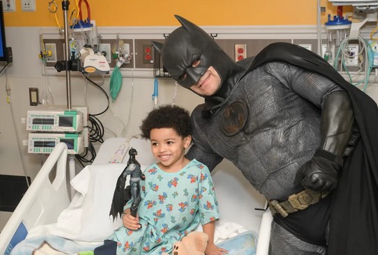 A person in a Batman costume strikes a heroic pose with a child clutching a Batman figure in the hospital room, brightening the day with superhero smiles and shared courage.