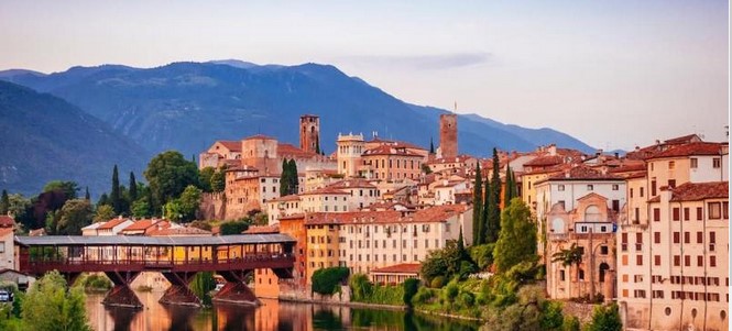Scenic view of Bassano del Grappa in Italy, featuring historic buildings and a wooden covered bridge over the Brenta River with mountains in the background, a testament to forgotten bravery woven through time.
