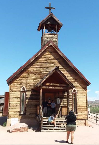 A small wooden chapel with a bell tower, set against the blue skies of Goldfield. Several people stand near the entrance and on the steps, their whispers mingling like ghostly echoes of the town's storied past.