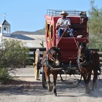 A person wearing a hat drives a red horse-drawn carriage with two horses on a dirt road, reminiscent of an iconic scene from the golden age of filmmaking, with a church visible in the background.