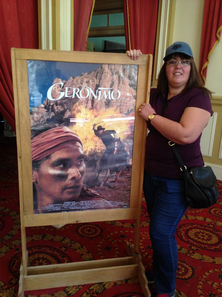 A woman in a cap stands next to a framed poster of a film titled "Geronimo," reflecting her passion for the film industry, in a room with red carpet and curtains.