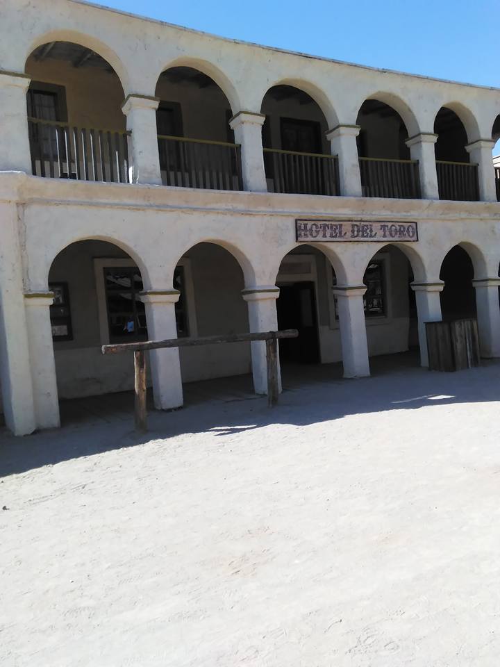 A two-story building labeled "Hotel Del Toro" proudly stands with its one elegant arched balcony, set against a sandy foreground under a clear blue sky.