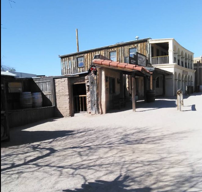 A dusty street scene with weathered wooden and stucco buildings, resembling an old western town under a clear blue sky, is enough to transport you back in time.