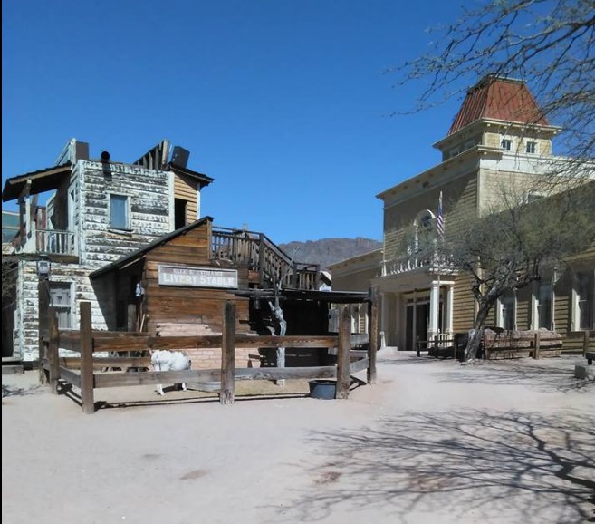 In a dusty Old West town scene, wooden buildings, including a livery stable, stand under a clear blue sky—evoking an era when one horse was enough for every cowboy.