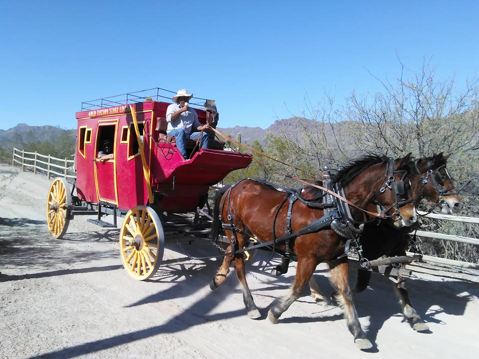 In a stunning continuation of the wild west tale, a red stagecoach pulled by two brown horses travels on a dirt road, driven by a person in a cowboy hat.