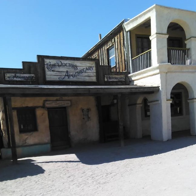 An old Western-style building with a sign reading "The Doctor's Apothecary" stands proudly under the clear blue sky. Its quaint wood and stucco elements, along with the inviting porch and upper balcony, are enough to transport one back in time.