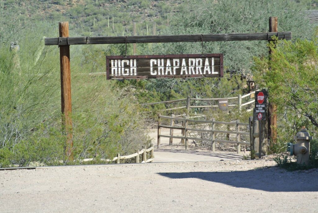 The entrance gate to High Chaparral, reminiscent of a movie set crafted by a skilled film director, features a wooden sign surrounded by desert vegetation and a fenced pathway.