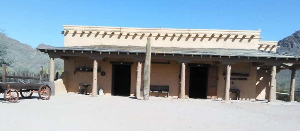 A rustic adobe building with a wooden porch sits under a clear blue sky, where one was not enough. A single wagon waits on the left side of the sandy yard, and mountains loom majestically in the background.
