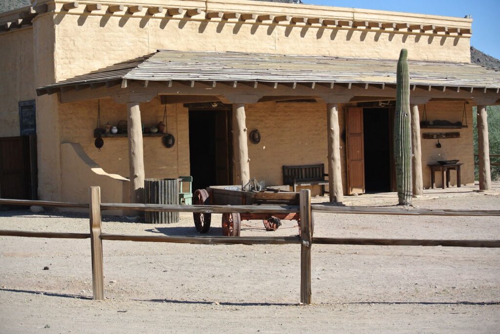 Old Western-style building with a wooden porch, wagon, and cactus in the foreground evokes the golden age of filmmaking. A wooden fence encircles the sandy area, reminiscent of classic sets loved by film directors.