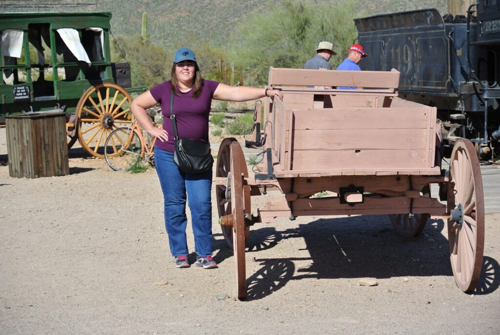 A woman in a purple shirt stands next to an old wooden wagon in a desert setting, as if she's scouting locations for her next filmmaking project, with a green carriage and cacti visible in the background.
