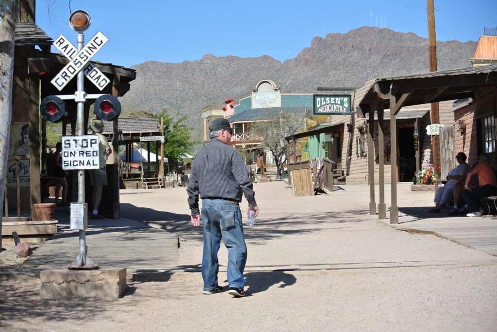 A film director strides through a western-themed town, with a railroad crossing and majestic mountains painting the backdrop.